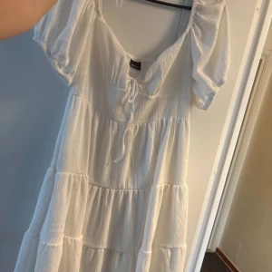 Vit klänning från Gina Tricot - Säljer en söt vit klänning från Gina Tricot med korta ärmar och knytning framtill. Klänningen har en luftig och bekväm passform med flera volanger som ger en fin rörelse. Perfekt för varma sommardagar! 🌞