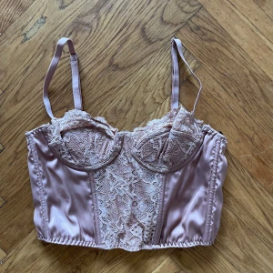 Korsettopp med spetsdetaljer - Rosa/rostbrun korsettopp med snygga spetsdetaljer och justerbara axelband. Toppen har en satinliknande finish och hakknäppning bak. 