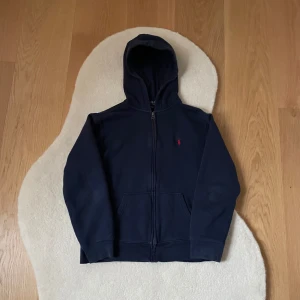 Mörkblå hoodie från Polo Ralph Lauren - Säljer en snygg mörkblå hoodie från Polo Ralph Lauren med dragkedja och klassisk logga i rött på bröstet. Perfekt för en avslappnad stil med sina långa ärmar och bekväma passform. Huvan ger extra värme och stil. Passar perfekt till vardagsoutfiten.