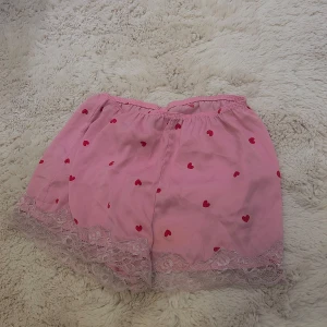 Rosa shorts med hjärtan och spetsdetaljer - Söta rosa shorts dekorerade med röda hjärtan och spetsdetaljer längs nederkanten. Perfekta för en mysig dag hemma eller som en del av en söt outfit.