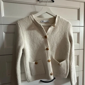 Beige kofta från Zara - Säljer en stilren beige kofta från Zara med dekorativa guldknappar och två praktiska fickor framtill. Koftan är stickad och har långa ärmar, perfekt för kyligare dagar. Ett måste i garderoben för en klassisk look.