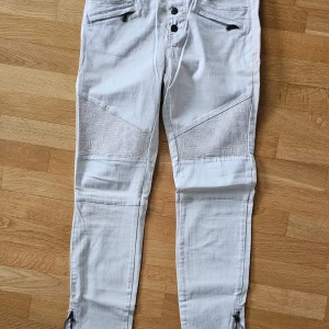 Beige jeansbyxor från KappAhl - Snygga beige jeansbyxor från KappAhl med unika detaljer. Byxorna har knappar framtill och förstärkta knän med textur. Kedjan på benen nertill på sidan. Midjehöjd ( lågmidjade) 23,5 innerbensmått 74 cm Midjan 39*2