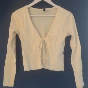 Cremevit kofta från H&M Divided  - Säljer en vit kofta från H&M Divided. Den har långa ärmar och en knytning framtill. Perfekt för lager-på-lager och att ha över en topp. Koftan är i ett mjukt material och har en lätt ribbad kant.