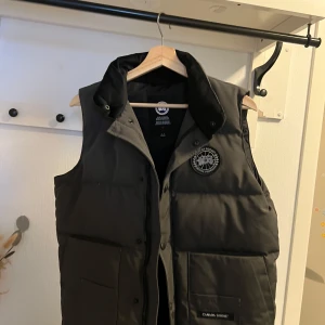 Canada Goose väst grå S  - Säljer en svart dunväst från Canada Goose med en broderad logga på bröstet. Västen har en dragkedja och knappar framtill samt praktiska fickor. Perfekt för kyligare dagar.INGA SKADOR! 