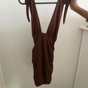 Brun klänning med djup urringning - Snygg brun klänning med djup urringning och smala axelband. Perfekt för en elegant look. Klänningen har en åtsittande passform och är tillverkad i ett mjukt material.