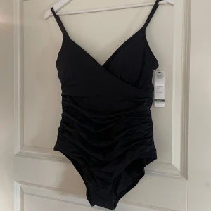 Svart baddräkt från Lindex - Snygg svart baddräkt från Lindex med omlott-design och justerbara axelband. Tillverkad i återvunnen polyamidblandning, perfekt för en stilren look vid poolen eller stranden. Räfflad framsida och slät baksida för en fin passform. Oanvänd med hygien klisterlappen och prislappen kvar. Kanske blir detta din nya baddräkt inför sommaren?