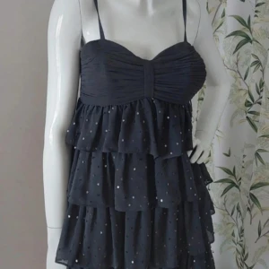 Svart glittrig klänning med volanger - Snygg svart klänning med smala axelband och flera lager av volanger. Klänningen har ett glittrigt mönster som ger en festlig känsla. Perfekt för en utekväll eller speciella tillfällen.