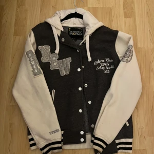 Grå och vit bomberjacka från Brave New World - Säljer en grå och vit bomberjacka från Brave New World med broderade detaljer och knappar framtill. Jackan har långa ärmar och ribbade muddar. Perfekt för en cool och avslappnad stil.