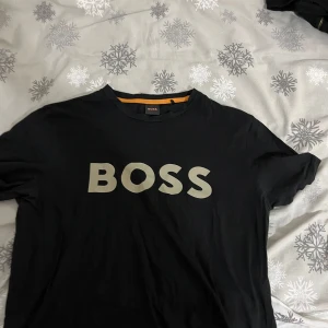 Svart t-shirt från Boss - Snygg svart t-shirt från Boss med stort tryck på framsidan. Perfekt för en stilren och avslappnad look. T-shirten har korta ärmar och en klassisk rund halsringning.Pris kan diskuteras!!