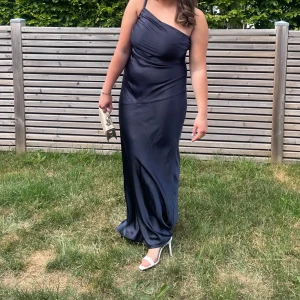 Mörkblå långklänning med oneshoulder - Elegant mörkblå långklänning i satin med en stilren oneshoulder-design. Perfekt för festliga tillfällen eller balen