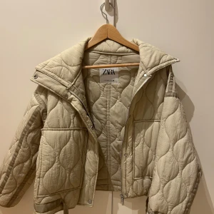 Beige quiltad jacka från Zara - Säljer en snygg beige quiltad jacka från Zara. Jackan har en dragkedja framtill och tryckknappar vid kragen. Den är nästintill oanvänd och i super bra skick!!