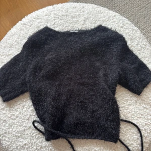Svart omlott tröja från NA-KD - Säljer en svart, fluffig omlott tröja från NA-KD designad av Hanna Schönberg. Tröjan har korta ärmar och knytband i midjan för en justerbar passform. Perfekt för en stilren och mysig look.