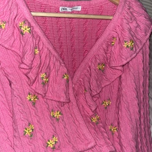Rosa stickad tröja från Zara - Säljer en söt rosa stickad tröja från Zara med broderade blommor och volangdetaljer. Tröjan har knappar framtill och en omlott-design. Perfekt för en färgglad look! Bara använt den två eller tre gånger ;)