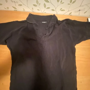 Svart pikétröja från Clique - Säljer en klassisk svart pikétröja från Clique. Tröjan har en krage och knappar framtill, vilket ger en stilren look. Perfekt för en avslappnad men ändå snygg stil. Pris kan diskuteras vid snabb affär