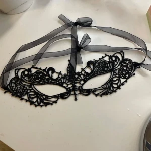 Svart spetsmask - Elegant svart mask i spets med intrikata detaljer. Perfekt för maskerad eller festliga tillfällen. Den har ett band för att knyta runt huvudet, vilket ger en justerbar passform.