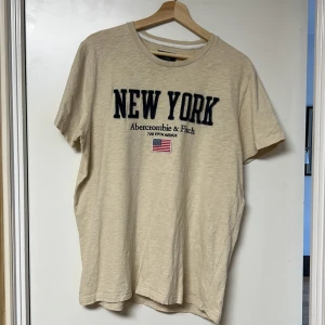 Beige t-shirt från Abercrombie & Fitch - Säljer en beige t-shirt från Abercrombie & Fitch med texten 'New York' och en liten amerikansk flagga på framsidan. T-shirten har en rund hals och korta ärmar. Perfekt för en avslappnad stil.