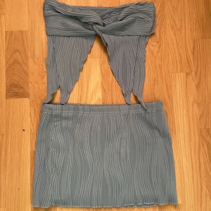 Grå axelbandslös klänning - Snygg grå axelbandslös klänning med en unik twistdetalj framtill. Klänningen har en ribbad struktur och en kort längd som ger en modern look. Perfekt för en stilren och chic outfit.