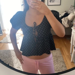 Svart blus med vita prickar - Säljer en söt svart blus med vita prickar och puffiga korta ärmar. Blusen har en knytning framtill och en lätt volang vid nederkanten. Perfekt för en somrig look!