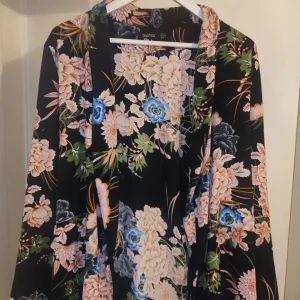 Blommig kimono från Boohoo - Säljer en snygg blommig kimono från Boohoo. Den har ett färgglatt blommönster i rosa, blått och grönt på en svart bakgrund. Kimonon har långa ärmar och ett knytband i midjan. Perfekt för att lägga till en färgklick i garderoben!