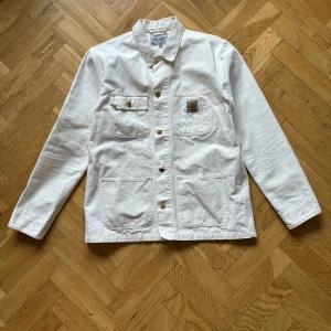 Carhartt Michigan coat vit  - Säljer en stilren vit overshirt från Carhartt med knappar framtill och flera praktiska fickor. Perfekt för lager-på-lager-stilen. Den har långa ärmar och en klassisk krage. Enkelt att matcha med olika outfits. Storlek XL