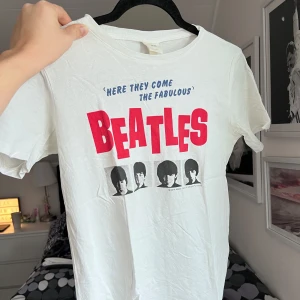 Vit Beatles t-shirt - Säljer en vit t-shirt med tryck av The Beatles. Texten 'Here they come the fabulous Beatles' i blått och rött pryder framsidan tillsammans med svartvita porträtt av bandmedlemmarna. Perfekt för alla Beatles-fans! 🎸 Storlek: XS. Axel: 38cm. Längd: 50cm.