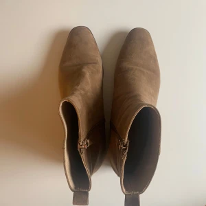 Bruna mocka boots med dragkedja - Snygga bruna/beige/sandfärgade boots i fuskmocka (OBS ej äkta mocka) med dragkedja på sidan. De har en spetsig tå och låg klack, vilket ger en stilren look. Perfekta för höst och vår. Köpta på HM, knappt använda. Frakt tillkommer på priset