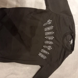 TRAPSTAR SWEATER - Säljer en svart långärmad tröja från Thrasher med vit text i gotisk stil på bröstet. Tröjan har en rund halsringning och är tillverkad i ett mjukt material. Perfekt för en avslappnad stil.