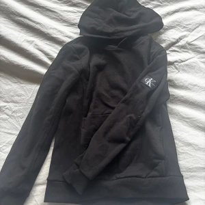 Svart hoodie från Calvin Klein Jeans - Snygg svart hoodie från Calvin Klein Jeans med en klassisk design. Den har en stor huva och en praktisk magficka. Perfekt för en avslappnad stil. Vi säljer den på grund av att den är för liten. Ny skick, använd nån gång. Storleken är 12 år, motsvarar ungefär stl 152.