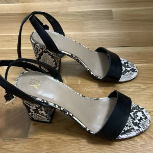 Klackar med ormmönster - Snygga klackar från XIT med en svart rem över foten och ormmönstrad sula och klack. Nästan helt oanvänd