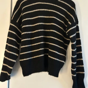 Svart stickad tröja  - Säljer en svart stickad tröja med beiga ränder. Tröjan har långa ärmar och en bekväm passform. 