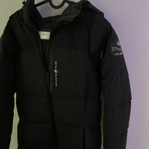 Svart dunjacka från Sail Racing - Snygg svart dunjacka från Sail Racing med huva och dragkedja. Jackan har logotypdetaljer på ärmen och bröstet. Perfekt för kalla dagar med sin varma och skyddande design.