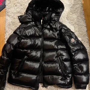 Moncler maya - Säljer en snygg svart pufferjacka från Moncler. Den är perfekt för kalla dagar med sitt blanka material och tjocka vaddering. Jackan har en dragkedja framtill och en mysig huva. Insidan har ett coolt serietidningsmönster. Perfekt för vintern! 🖤