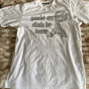 Vit t-shirt med tryck från Blinglucid - Säljer en vit t-shirt från Blinglucid i storlek S. Säljer den pga att jag köpte fel storlek. Den har ett coolt tryck med texten 'paris au clair de lune' och en bild av Eiffeltornet under en måne. Perfekt för en avslappnad stil eller en dag ute i solen. T-shirten är i bomull och har korta ärmar. Använd 2 gånger. Nypris:500kr
