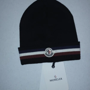 Svart mössa från Moncler - Säljer en snygg svart mössa från Moncler med klassiska ränder i vitt, blått och rött. Den är i nyskick och har en broderad Moncler-logga framtill. Perfekt för att hålla sig varm och stilren under kyliga dagar. Passar både till vardags och mer uppklädda tillfällen.