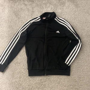 Svart träningsjacka från Adidas - Säljer en svart träningsjacka från Adidas i storlek 9-11y Jackan har de klassiska tre vita ränderna längs ärmarna och en dragkedja framtill. Perfekt för träning eller som en sportig vardagslook. Den är i bra skick och redo för nya äventyr! 🖤