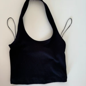 Svart ribbad topp från H&M Divided - Snygg svart ribbad topp från H&M Divided i storlek XS. Perfekt för sommardagar eller en utekväll. Den har en stilren design med smala axelband och en croppad passform. Toppen är i bra skick!