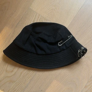 Svart bucket hat med säkerhetsnålar - Säljer en cool svart bucket hat med unika detaljer som säkerhetsnålar och ringar 💗 Hatten är i bra skick 