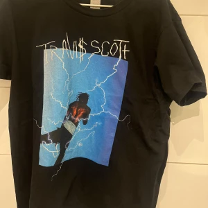 Svart t-shirt med Travis Scott-tryck - Cool svart t-shirt med ett stort tryck av Travis Scott på framsidan. Trycket har en blå bakgrund med blixtar och en siluett av artisten. Perfekt för fans av Travis Scott och streetwear-stil