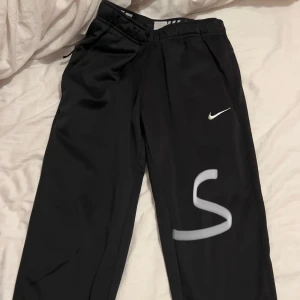 Svarta träningsbyxor från Nike - Säljer ett par svarta träningsbyxor från Nike i storlek S. Byxorna har en elastisk midja och är gjorda i ett mjukt och bekvämt material, perfekt för träning eller chill. De har en liten vit Nike-logga på ena benet. Passar både för gymmet och hemmamys!