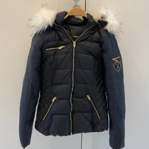 ROCKANDBLUE jacka - Snygg svart dunjacka från ROCKANDBLUE med quiltat mönster och vita pälsdetaljer på huvan. Jackan har flera praktiska dragkedjor i guld och en logga på ärmen. Perfekt för kalla vintrar.