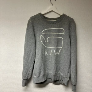 Grå sweatshirt från G-Star RAW - Vintage sweatshirt från G-Star RAW. Den har ett stort tryck på framsidan med texten 'RAW'. Säljs inte i butik längre. Jätteskön. Märkeströja. Bra skick 