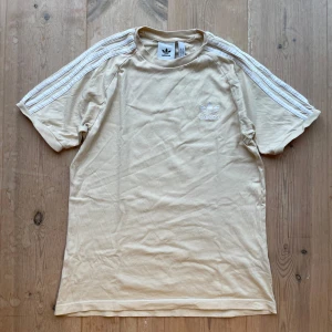 Beige t-shirt från Adidas - Säljer en beige t-shirt från Adidas med klassiska tre ränder på ärmarna och broderad logga på bröstet. T-shirten är kortärmad och tillverkad i bomull. Skriv vid frågor!