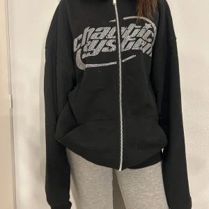 Svart hoodie med dragkedja - Säljer en svart hoodie med dragkedja och ett stort tryck i silver på framsidan. Den har en bekväm passform och en praktisk huva. Perfekt för en avslappnad stil.
