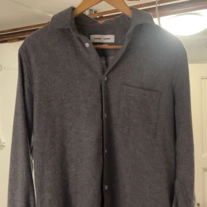 Grå flanel skjorta från Samsoe Samsoe - Snygg grå flanel skjorta från Samsoe Samsoe med klassisk krage och knappar framtill. Skjortan har en bröstficka och är perfekt för en stilren look. Passar bra till både vardag och mer uppklädda tillfällen. (Den lägsta knappen på tröjan saknas, men annars felfri)