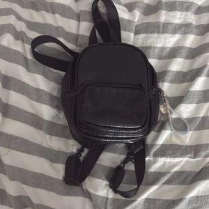Svart ryggsäck från Zara - Säljer en snygg svart ryggsäck från Zara med krokodilmönster. Väskan har en praktisk dragkedja och en cool nyckelring med glitterdetaljer. Perfekt för att bära dina dagliga nödvändigheter med stil.