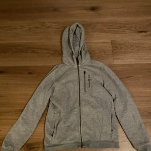 Grå hoodie från Sail Racing - Säljer en grå hoodie från Sail Racing med dragkedja och två fickor framtill. Tröjan har en justerbar huva och är perfekt för en avslappnad stil. Den är tillverkad i ett mjukt material som ger en bekväm känsla.