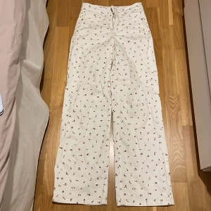 Vita blommiga jeans med dragkedja - Snygga vita jeans med ett sött blommönster i rosa och rött. Byxorna har en hög midja och en rak passform. De är utrustade med en dragkedja och knapp framtill. Storlek 34 🌸