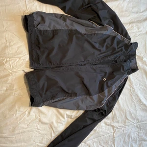 Svart och grå vindjacka från SOC - Säljer en svart och grå vindjacka från SOC i storlek XL. Jackan har en dragkedja framtill och är tillverkad i polyester. Perfekt för blåsiga dagar med sin lätta och funktionella design. OBS!! Ej funktionell dragkedja. 