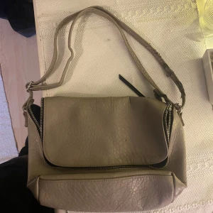 Beige axelväska med dragkedja - Snygg beige axelväska i skinn med praktiska dragkedjor. Perfekt för att bära dina dagliga nödvändigheter med stil. Axelremmen är justerbar för bästa passform.