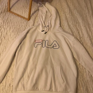 Vit hoodie från Fila - Säljer en vit hoodie från Fila med deras ikoniska logga i rött och blått på bröstet. Tröjan har en klassisk känguruficka och justerbar huva med snören. Perfekt för en avslappnad stil.